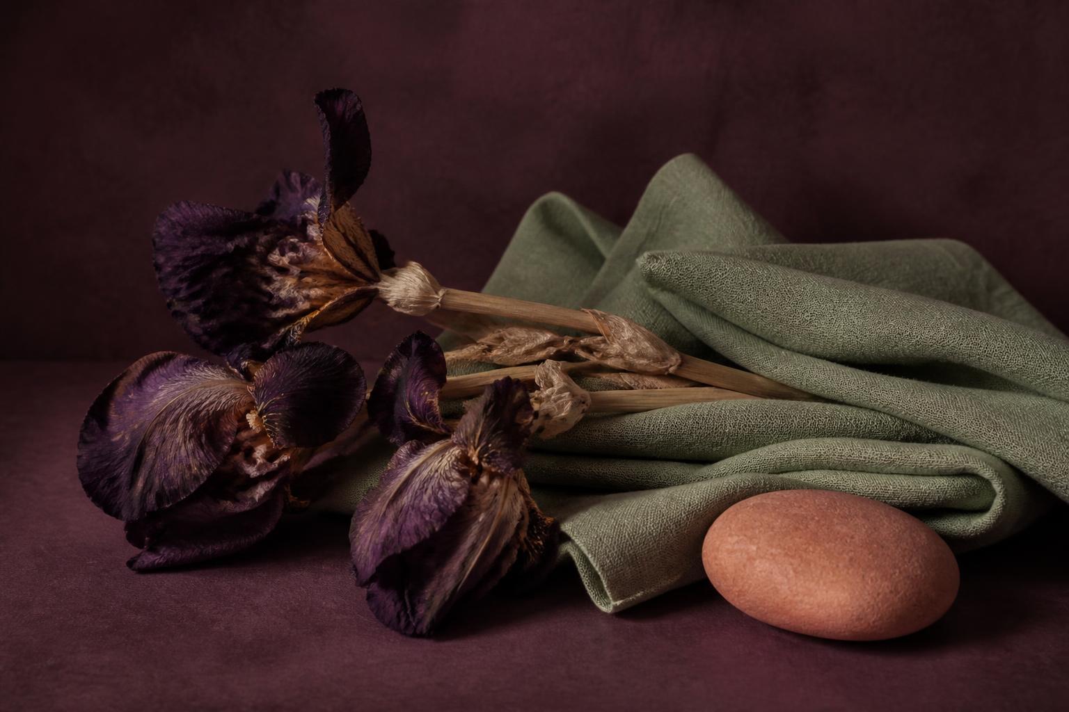 A still life of dried iris stems, folded sage-green linen and a smooth clay stone arranged on a deep plum surface — the calm, tactile rituals Emmenia suggests during a period